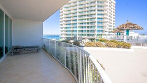 A balcony with a view of a tall hotel building, a thatched umbrella, and a body of water.
