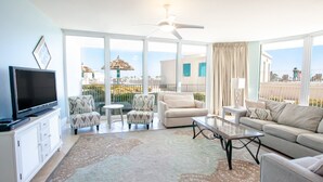 A spacious living room with a flat-screen TV, two sofas, a coffee table, and a ceiling fan. Large windows offer a view of the ocean and a building with a thatched roof.