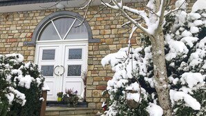 Une maison en pierre avec une porte et des fenêtres blanches, entourée d’arbres et de plantes couverts de neige.