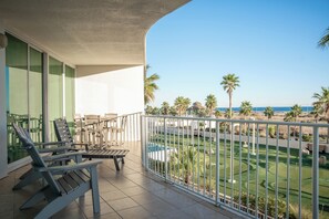 A balcony with two Adirondack chairs, a small table, and a view of a golf course with palm trees and a pool. - Three Bedroom Family-Friendly Condo Overlooking the Putting Greens. Complimentary Boat Slip! (Orange Beach)