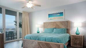 Un dormitorio con una cama grande, cabecera de madera, dos mesitas de noche con lámparas, un cuadro con una escena de playa y una puerta corrediza de vidrio que da a un balcón.