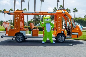 Ein Maskottchen in einem grünen Kostüm steht neben einem orangefarbenen Feuerwehrwagen.