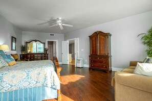 Une chambre spacieuse avec un plancher en bois, un grand lit recouvert d’une courtepointe à motifs floraux, une commode en bois, un fauteuil beige et un ventilateur de plafond.