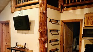 Een rustiek interieur met een houten zuil, een wandgemonteerde televisie, een houten tafel met spullen en een deur naar een andere kamer.