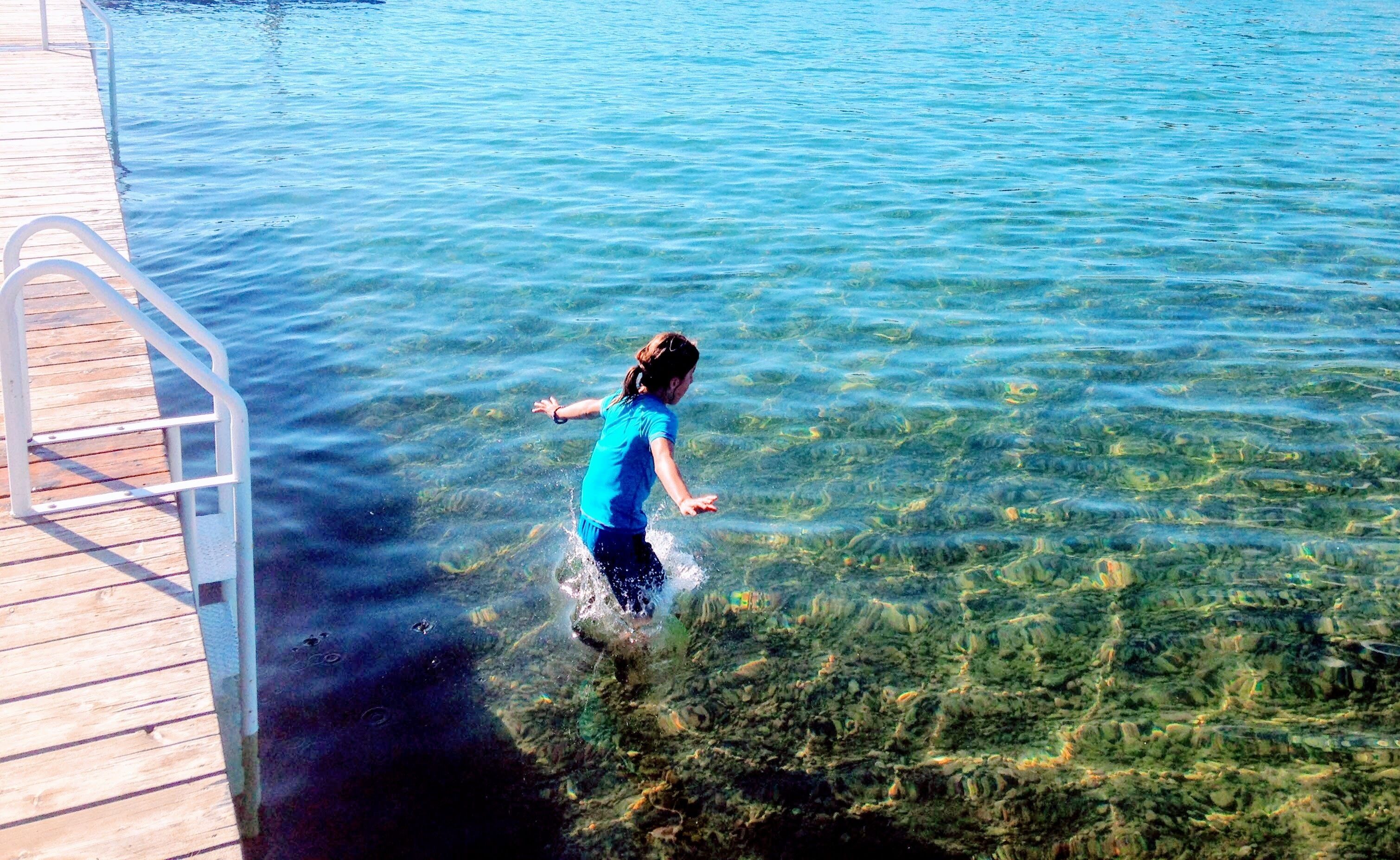 A person in a blue shirt wading in clear water near a wooden dock.
