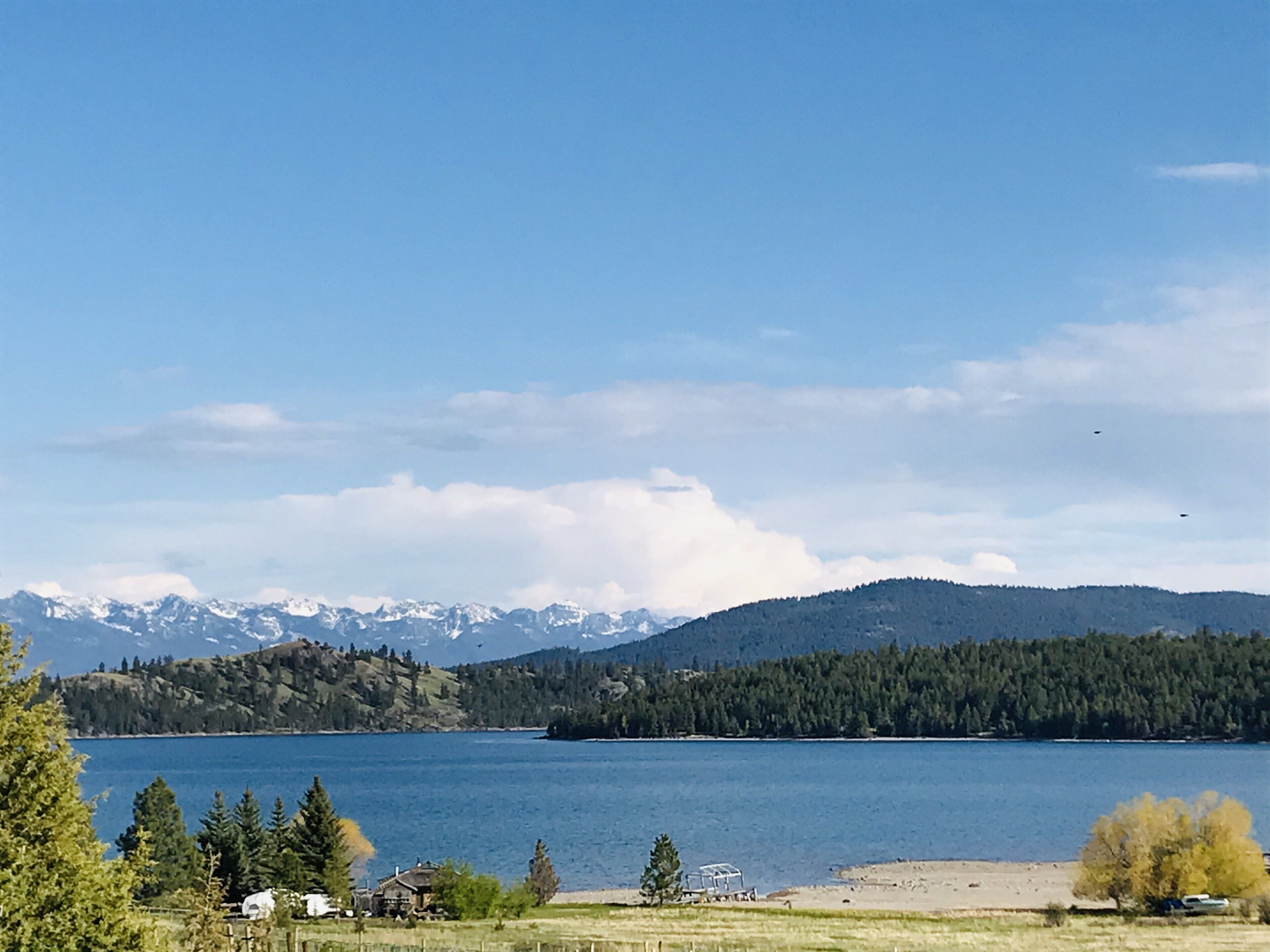 Um lago com montanhas ao fundo, rodeado por árvores e alguns edifícios.