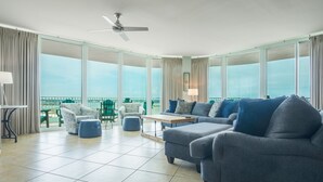 Ein geräumiges Wohnzimmer mit einer großen Sofagarnitur, einem Couchtisch und einem Deckenventilator. Große Fenster mit Vorhängen und ein Balkon mit Stühlen sind durch die Fenster sichtbar.