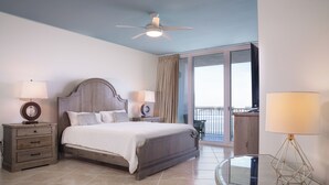 Une chambre à coucher comprenant un lit, deux tables de chevet avec des lampes, un ventilateur de plafond, une table en verre et une vue sur l’extérieur.