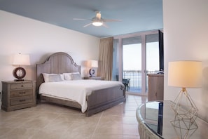 Un dormitorio con cama, dos mesitas de noche con lámparas, ventilador de techo, mesa de vidrio y vistas al exterior.