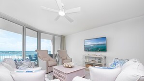 Una sala de estar moderna con un ventanal grande, ventilador de techo, televisor de pantalla plana y vista al océano.