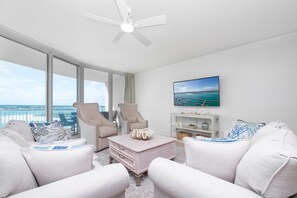 Un salon moderne doté d’une grande fenêtre, d’un ventilateur de plafond, d’un téléviseur à écran plat et offrant une vue sur l’océan.