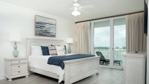 Ein Schlafzimmer mit Bett, Nachttischen, Deckenventilator, Gemälde und Blick auf den Balkon.