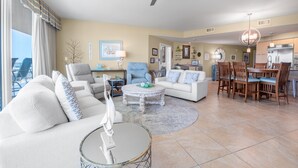 A spacious living area with a large window, a ceiling fan, a dining area with wooden chairs, and a kitchen in the background. - Condo with Panoramic Views of Gulf of Mexico. Complimentary Boat Slip! (Orange Beach)