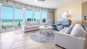 A spacious living room with a large window, a ceiling fan, and a round coffee table. - Condo with Panoramic Views of Gulf of Mexico. Complimentary Boat Slip! (Orange Beach)