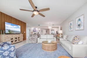 Ein geräumiges Wohnzimmer mit Deckenventilator, einem großen Flachbildfernseher, einem weißen Sofa, einem Korbtisch und einem gemusterten Ottoman.