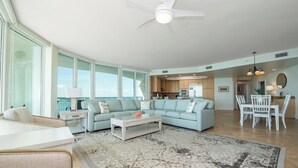 A spacious living room with a large L-shaped sofa, a coffee table, and a dining area with a white table and chairs. - Beautiful Family-Friendly Corner Condo with Panoramic Views! Complimentary Boat Slip! (Orange Beach)