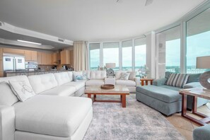 A modern living room with a large window, a kitchen in the background, and a view of the city. - Luxury Caribe Bay-View 4BR/3.5BA Condo (Orange Beach)