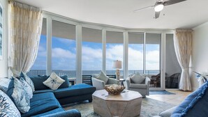 A spacious living area with a large window overlooking the sea, a ceiling fan, and a round coffee table. - Timeless Three Bedroom Penthouse with Gulf of Mexico Views (Orange Beach)