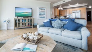 A living room with a sofa, a coffee table, a TV, and a kitchen in the background. - Timeless Three Bedroom Penthouse with Gulf of Mexico Views (Orange Beach)