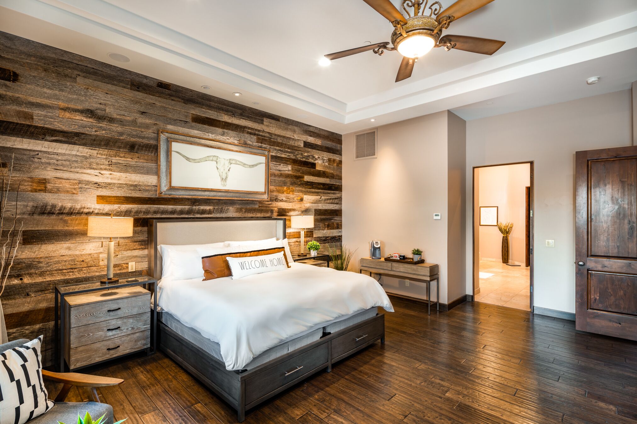Een slaapkamer met een groot bed, houten lambrisering, een plafondventilator en een nachtkastje met een lamp.