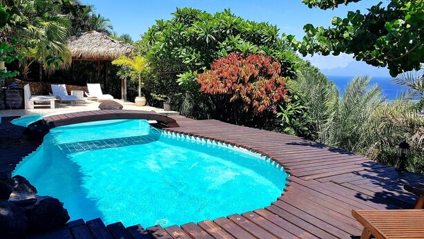 A swimming pool with a wooden deck, lounge chairs, and a thatched-roof structure.