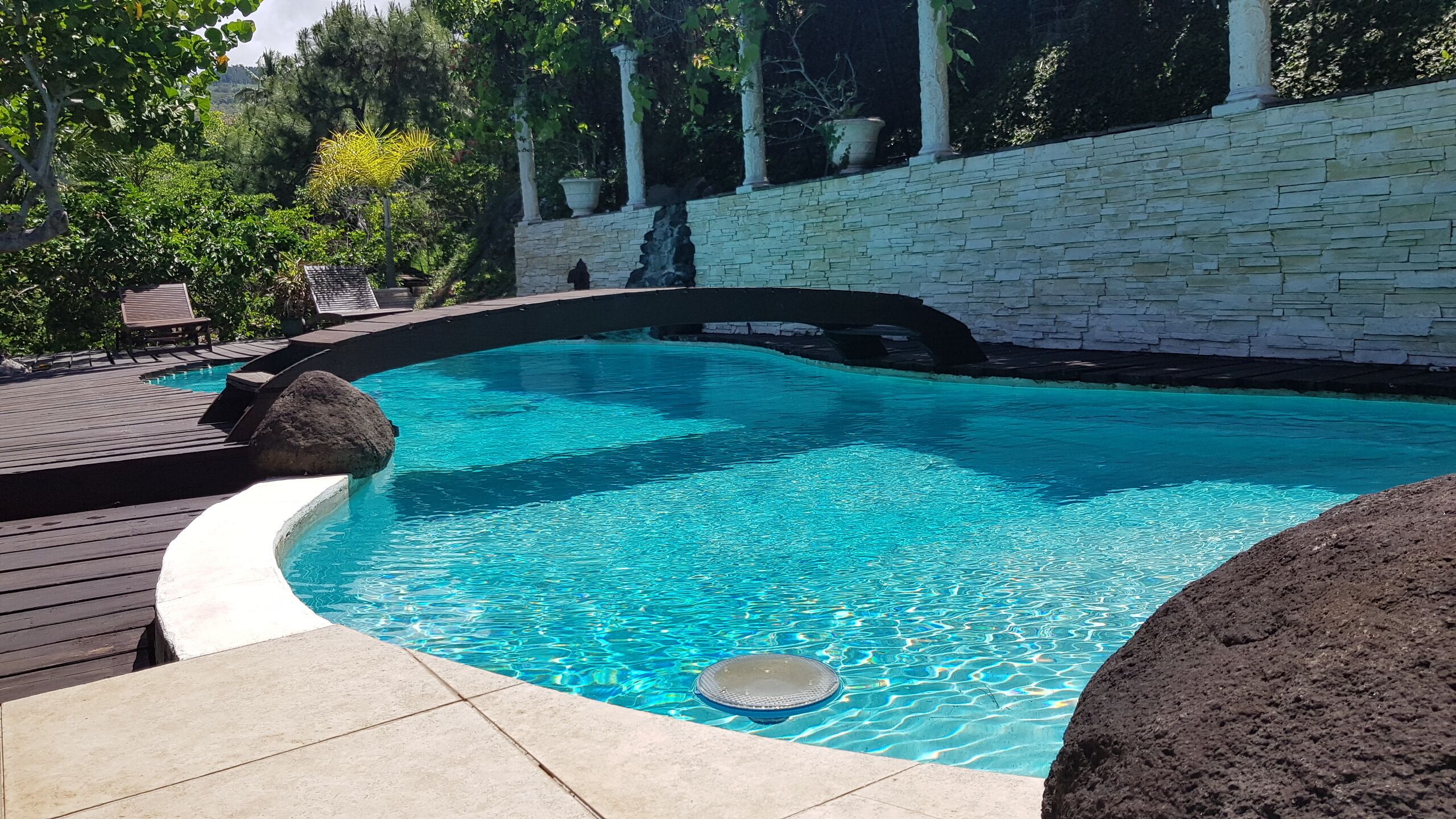 Une piscine avec un mur en pierre et une terrasse en bois.