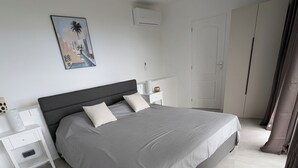A modern bedroom with a large bed, white nightstands, a wall-mounted air conditioner, and a door with a curtain.
