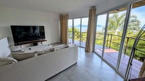 A modern living room with a large flat-screen TV on a white console table, a beige sofa, and a large sliding glass door leading to a balcony with a view of greenery and mountains.