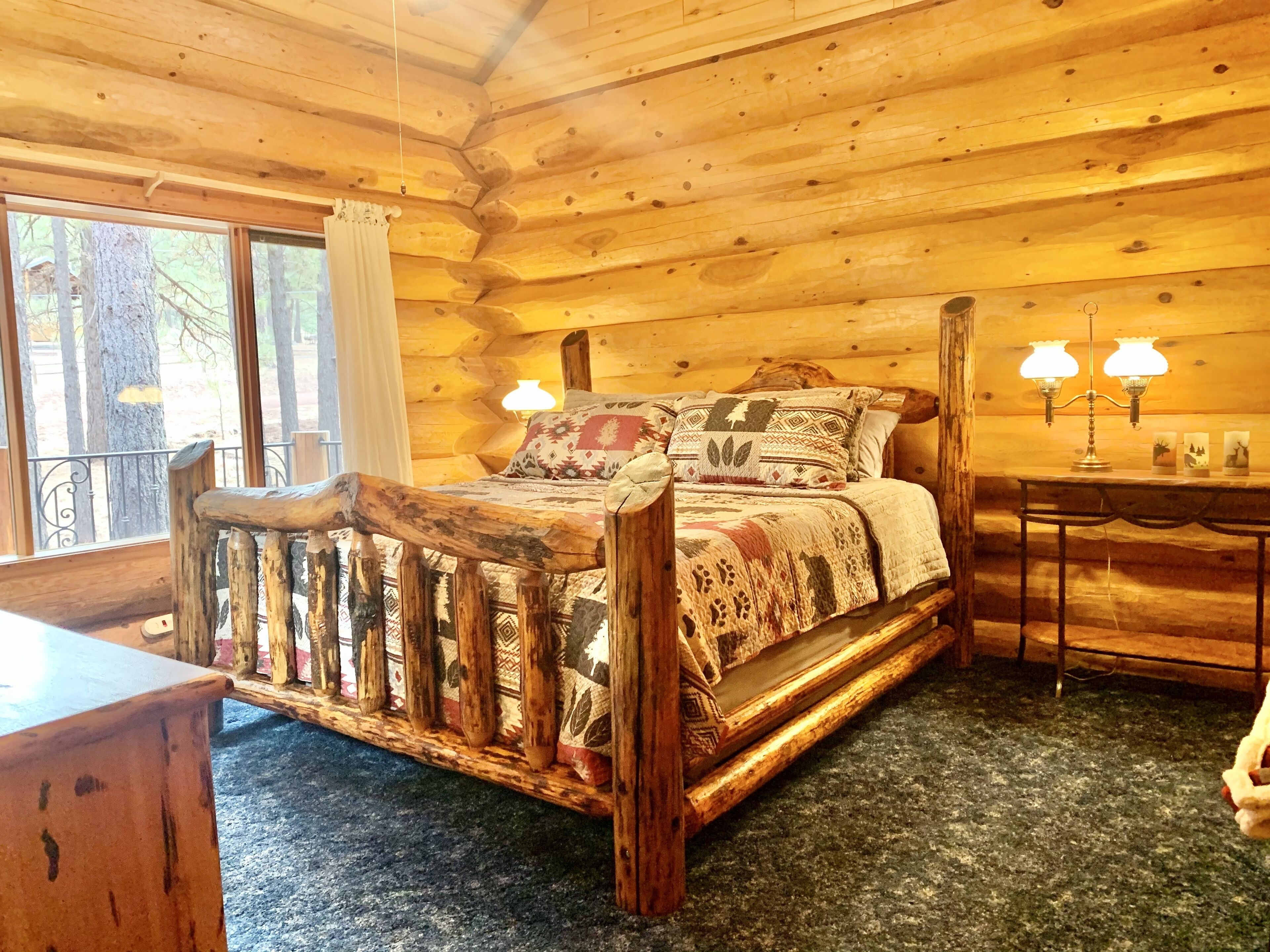 Une chambre située dans une cabane en rondins, avec un lit en bois, une petite table dotée de deux lampes et une vue sur les arbres à l’extérieur.