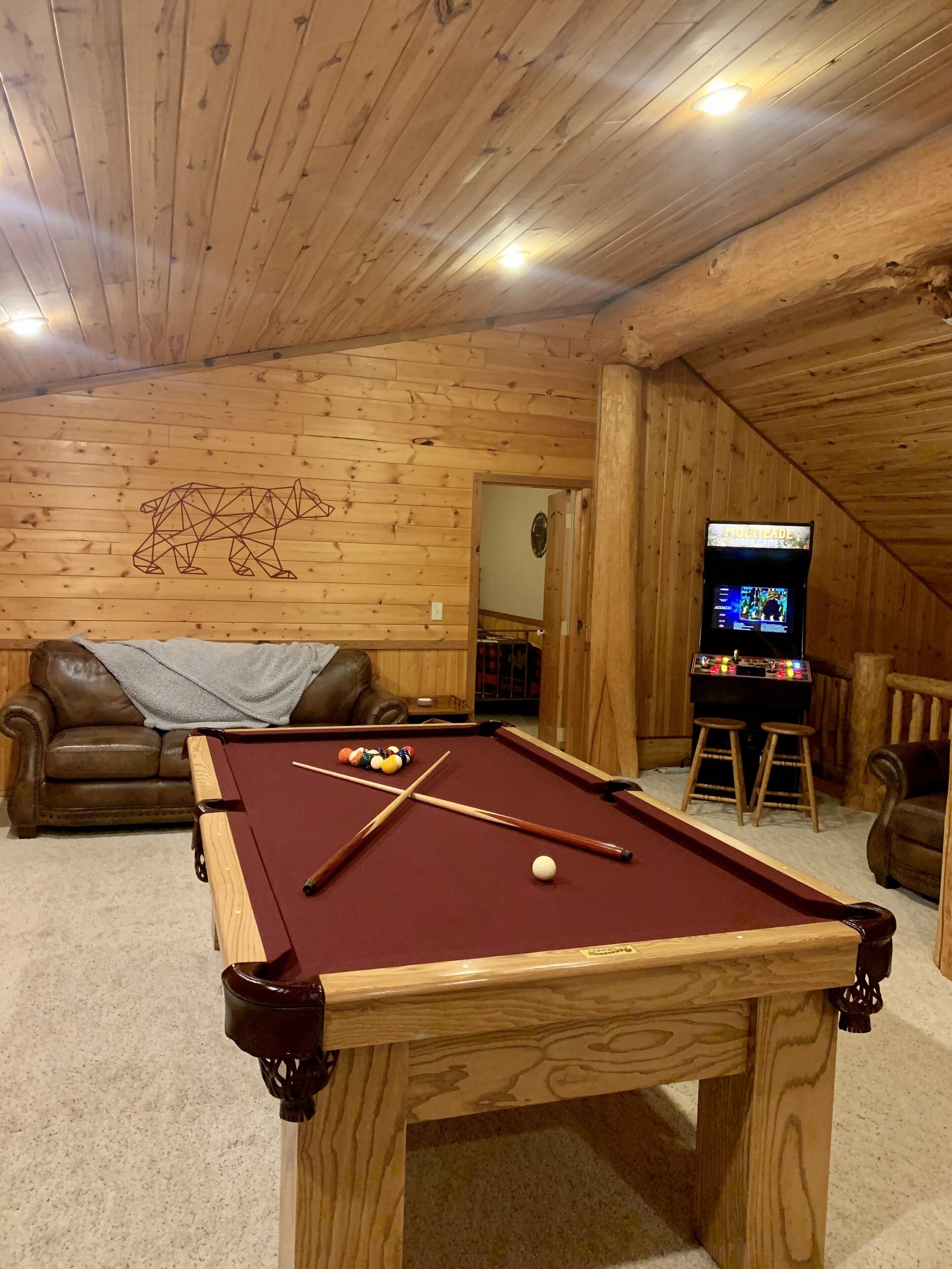 Une pièce aux murs lambrissés, avec une table de billard, un canapé en cuir marron et une console de jeux vidéo.