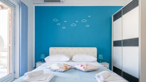 Un dormitorio con una pared azul, un cabecero blanco, dos almohadas blancas, una cama con colcha azul, dos almohadas de colores, una mesita de noche con lámpara y un armario.