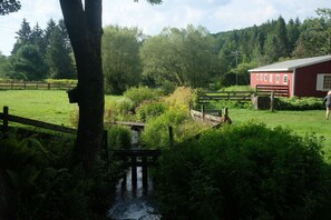 Paisaje rural con un pequeño arroyo, un puente de madera, un granero rojo y un cercado.