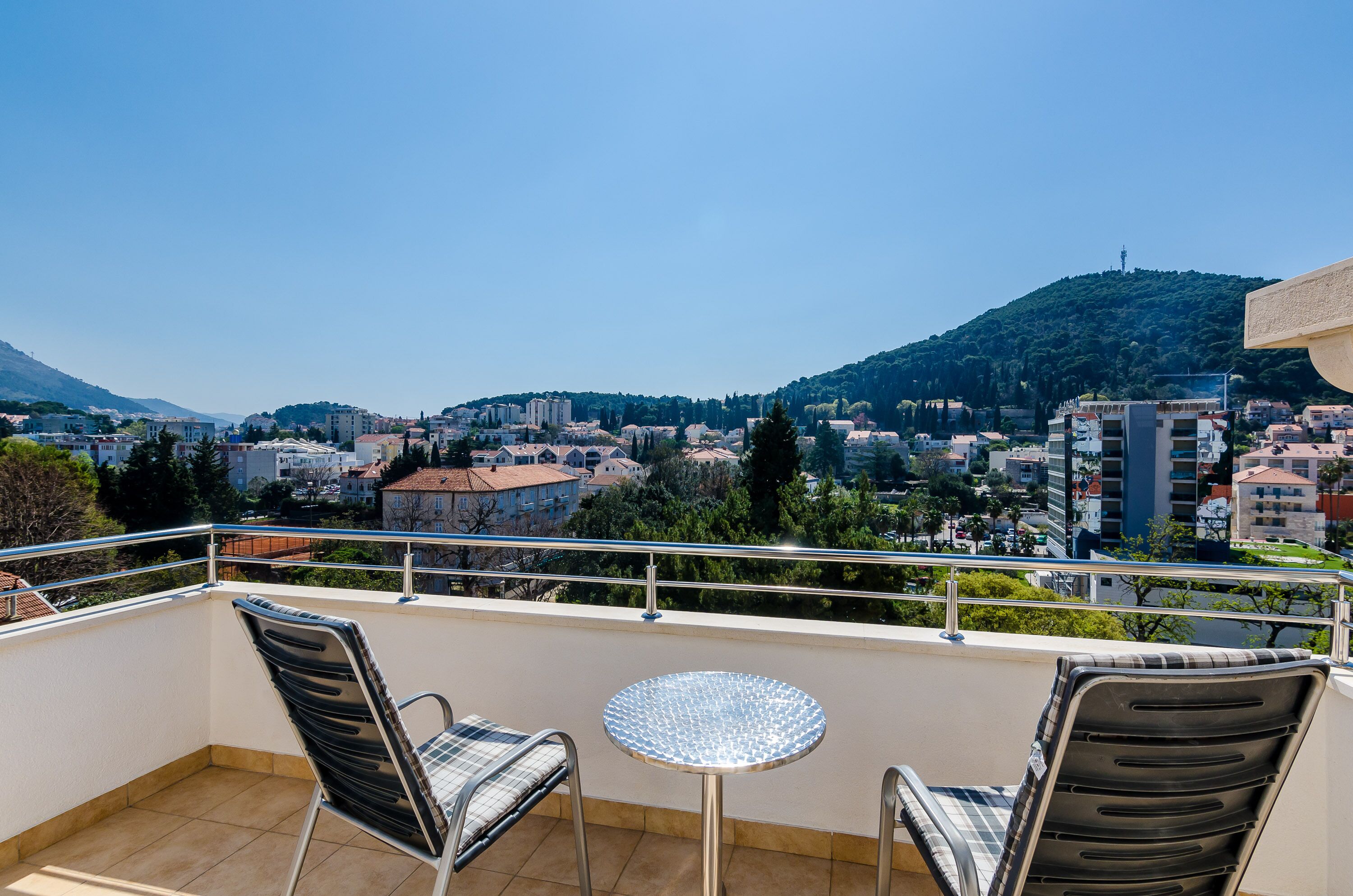 Un balcón con dos sillas y una mesa pequeña, con vista a un paisaje urbano que incluye edificios y una colina.