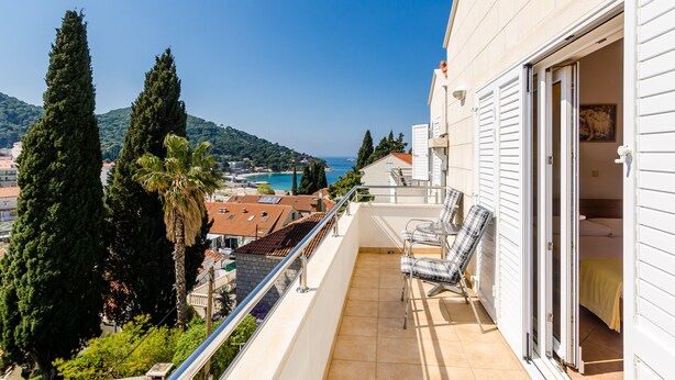 Un balcón con vistas al mar y montañas, una palmera, y un edificio con una puerta abierta.