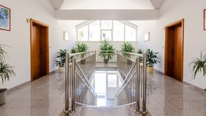 Un pasillo amplio y luminoso, con techo alto y ventanas grandes, que conduce a un rellano en el segundo piso con barandilla de metal.