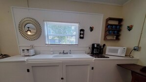 A compact kitchen with a white countertop, a sink, a window with blinds, a clock, and a coffee maker.