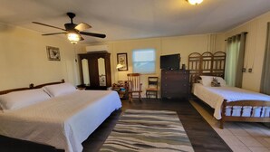 A hotel room with two beds, a ceiling fan, a desk with a television, and a large window with blinds.