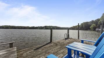 A wooden dock with blue Adirondack chairs, a metal ladder, and a view of a calm lake with trees in the background.