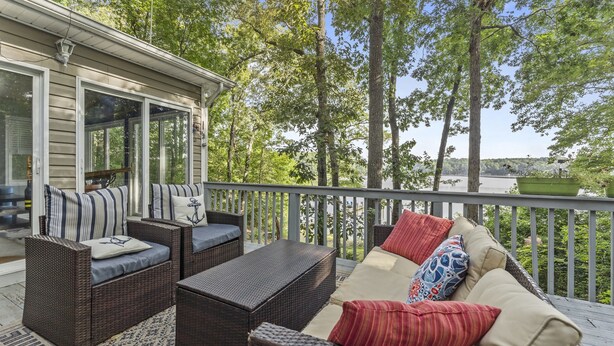 A covered outdoor deck with wicker furniture, a glass door leading inside, and a view of a lake surrounded by trees. - Waterfront 3BR/3BA Getaway: Beach, Dock, Firepit! AMAZING Sweeping VIEWS! (Heathsville)