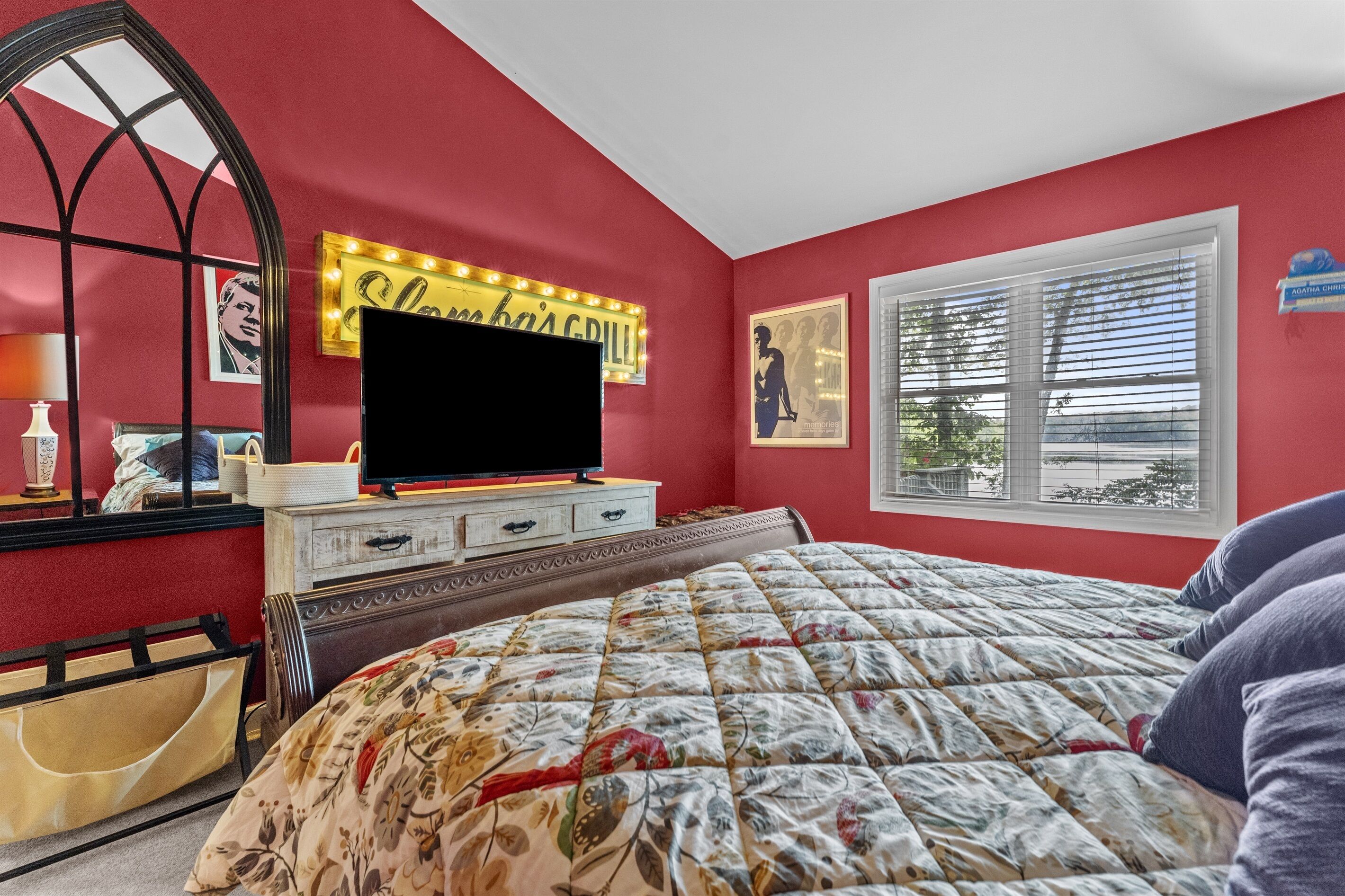 A bedroom with a red wall, a bed with a floral comforter, a TV on a wooden dresser, and a window with blinds.