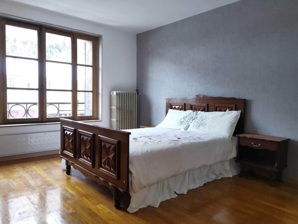 A bedroom with a wooden bed, a nightstand, a window with a view of buildings, and a radiator. - Appartement au bord de la Rivière (Modane)
