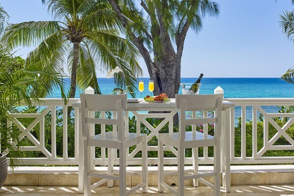 A white outdoor dining area with a table and chairs, overlooking a beach with palm trees and the ocean.