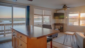 A spacious room with a large wooden table, a comfortable sofa, and a TV. It features a balcony with a view of the water and a sunset. - Sunset Views over Rachel Carson Preserve. Relax steps from Wells Beach. (Wells)