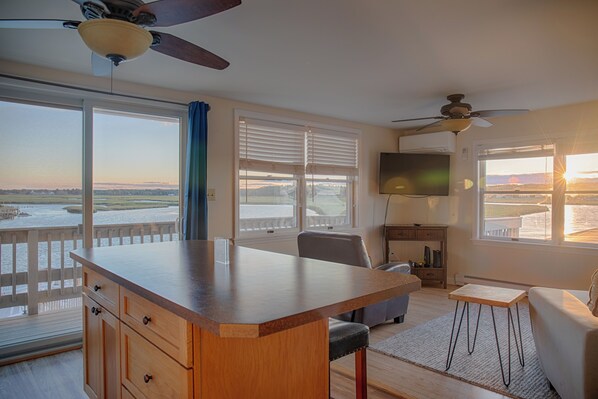 A spacious room with a large wooden table, a comfortable sofa, and a TV. It features a balcony with a view of the water and a sunset.
