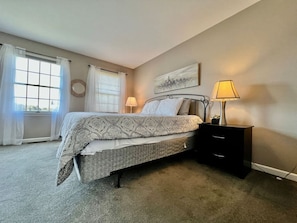 Une chambre à coucher comprenant un lit, des tables de chevet, une lampe, une fenêtre avec des rideaux et une œuvre d’art accrochée au mur.