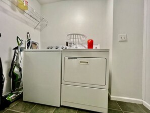A compact laundry room with a front-loading washer and dryer, a metal drying rack, and a vacuum cleaner. - 5042 Princeton Glend · Worley`s House (Liberty Township)
