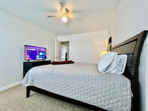 Quarto com cama, televisão, ventoinha no teto e porta para outro cômodo.