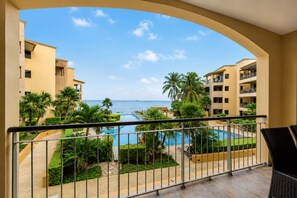 A balcony with a view of a swimming pool, palm trees, and a building with balconies.