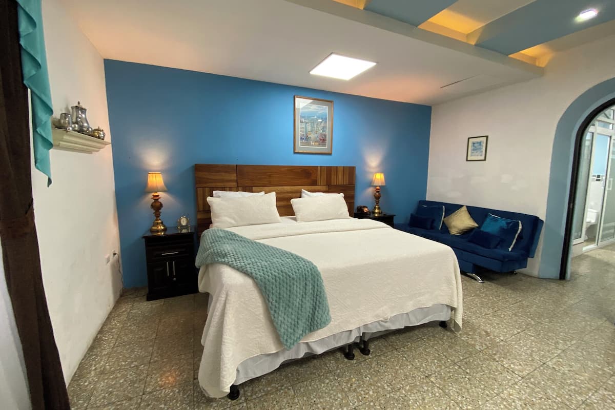 A bedroom with a large bed, two bedside tables with lamps, a blue wall, and a cityscape painting.