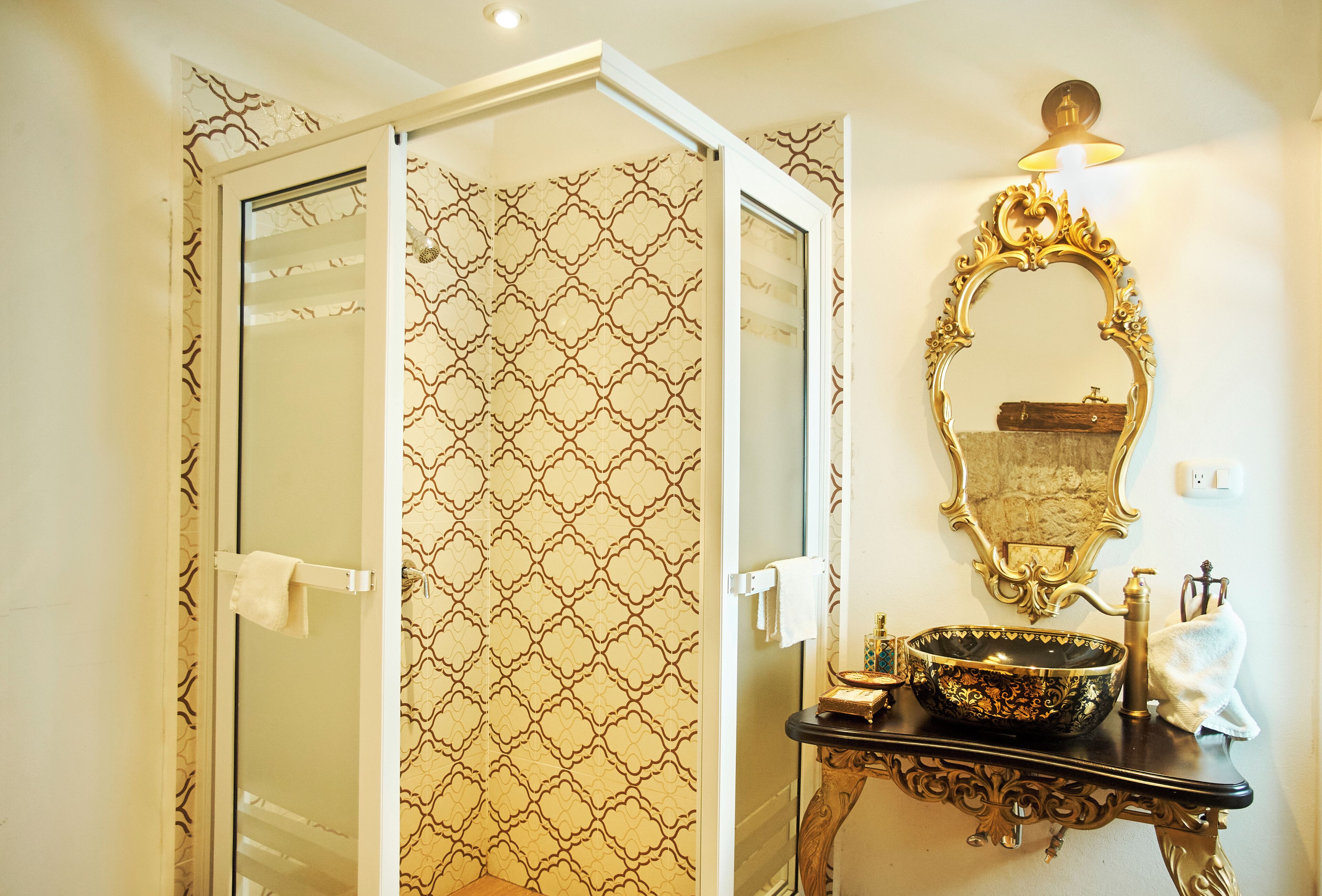 Ein Badezimmer mit gemustertem Wandpapier, einer Duschkabine aus Glas, einem spiegel mit goldfarbener Rahmung und einem schwarzen Waschbecken mit Dekorationen.